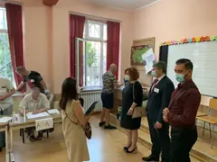 Наблюдатели с маски и баджове в избирателна секция в българско училище, членове на комисията работят на маса
